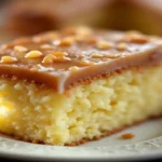 A close-up view of a slice of moist yellow kefir sheet cake topped with a smooth caramel glaze and sprinkled with chopped nuts, served on a white plate.